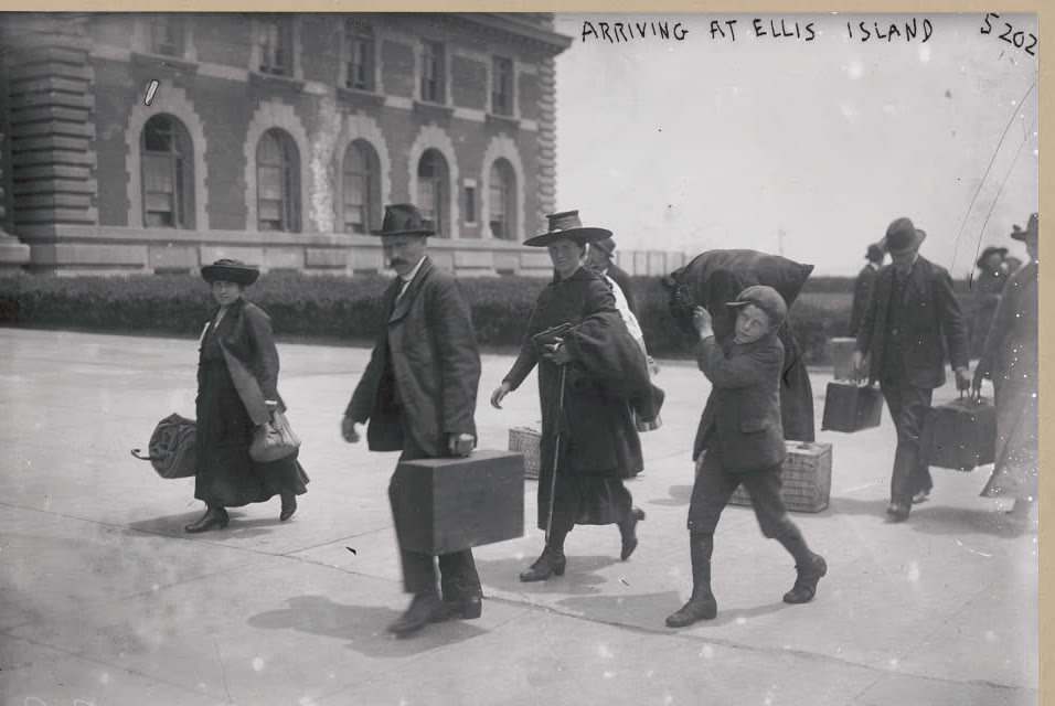 Immigrants carrying luggage walking outside the Ellis Island immigration building